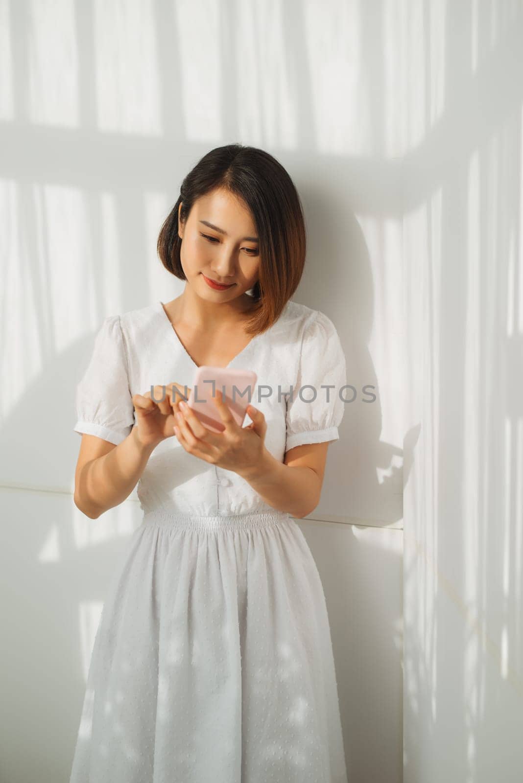 Young woman texting on her smartphone with a look of concentration as she stands against a white wall by makidotvn