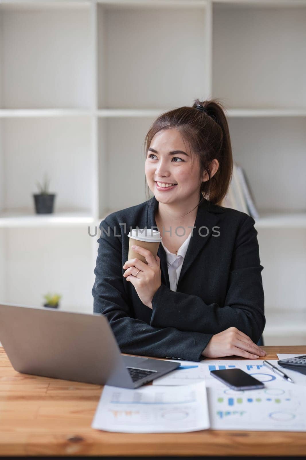Photo of positive Asian businesswoman holds a cup of coffee imagines plan holiday in office. by wichayada
