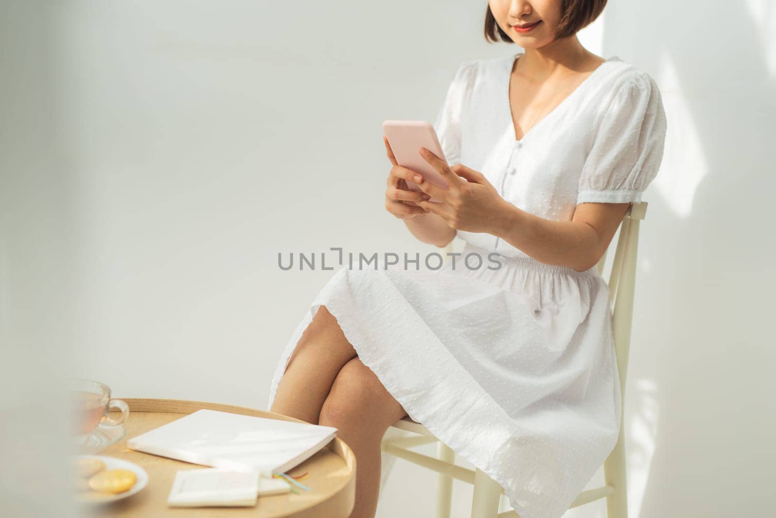 Beautiful young asian woman standing on white wall relax texting message on cellphone. by makidotvn
