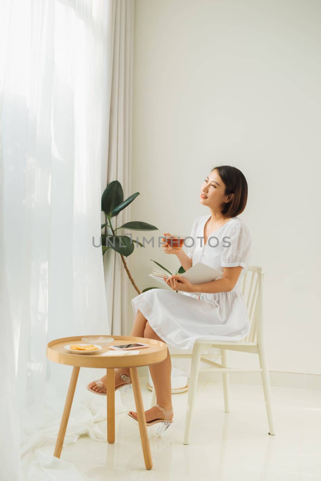 Young woman at home sitting near window relaxing in her living room reading book and drinking tea