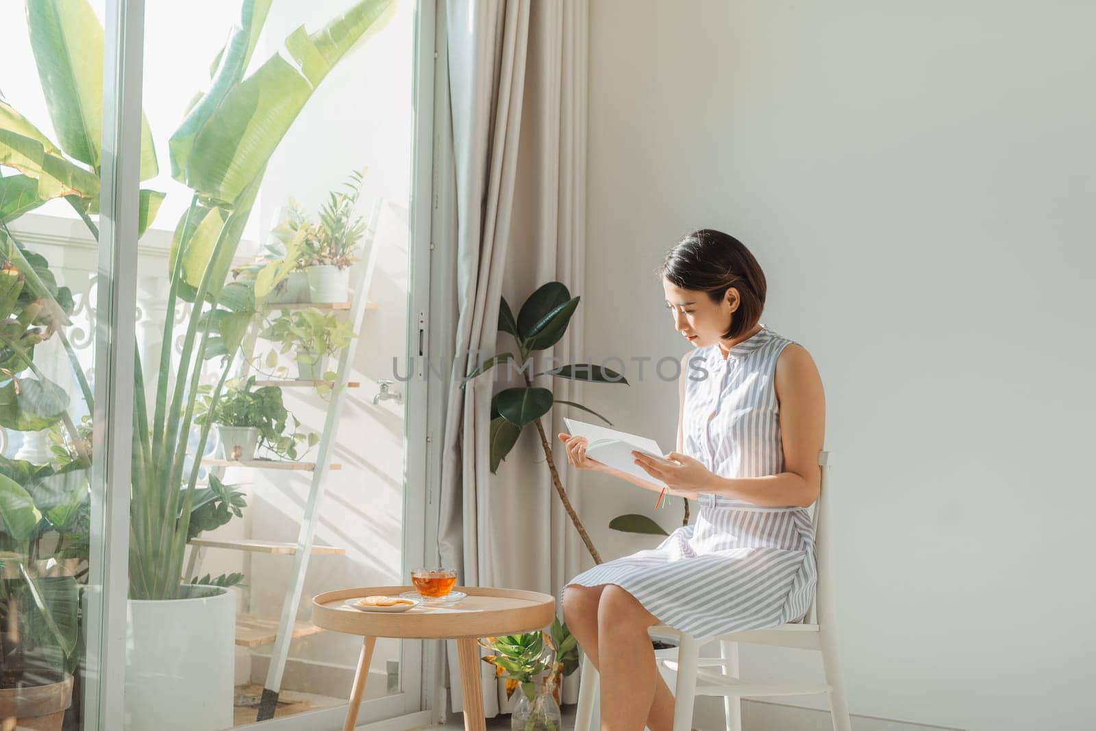 Young charming woman sitting on chair and reading book next to window at home.