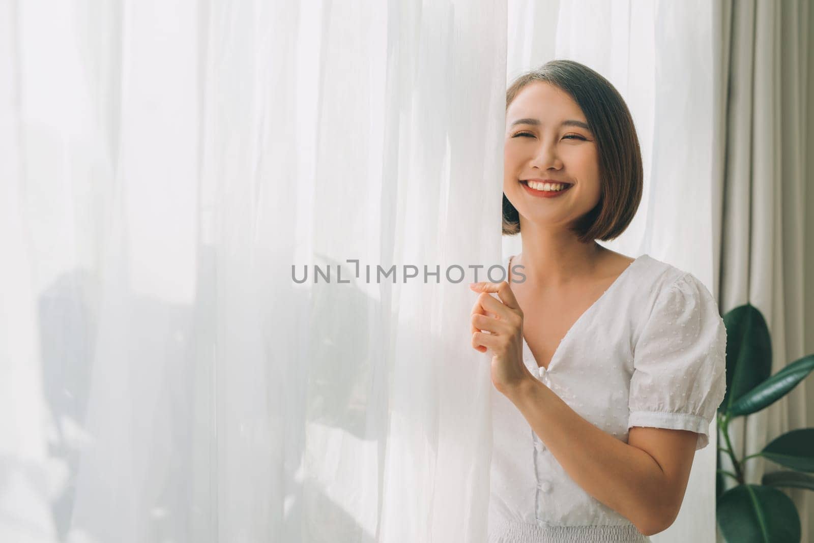 Side view of a happy woman opening curtains of a window and enjoying a new day with a warm light from outdoors by makidotvn