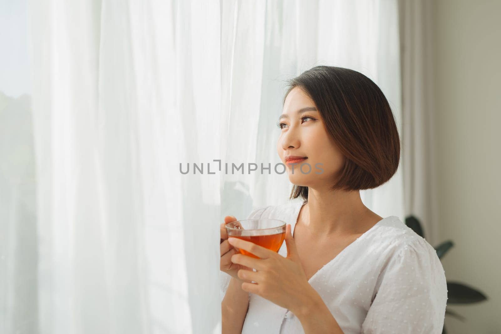 Portrait of a satisfied woman thinking looking at side holding a tea mug beside a window at home by makidotvn