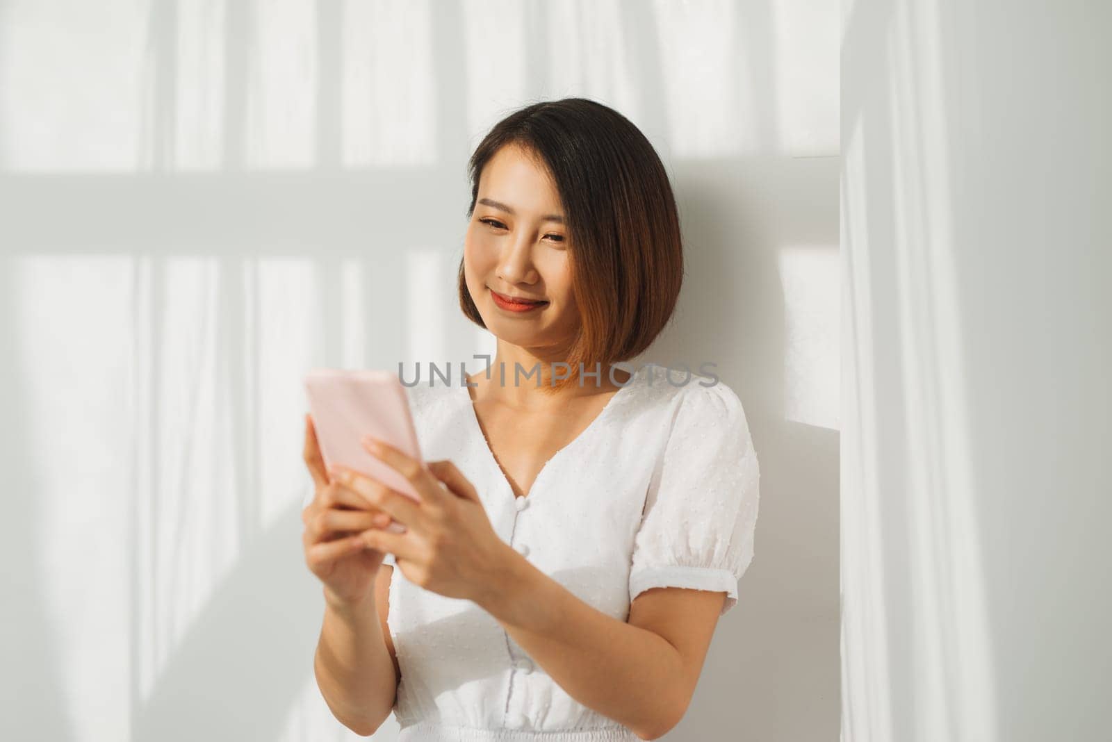 Content woman chatting on mobile phone while standing against white wall in sunset light