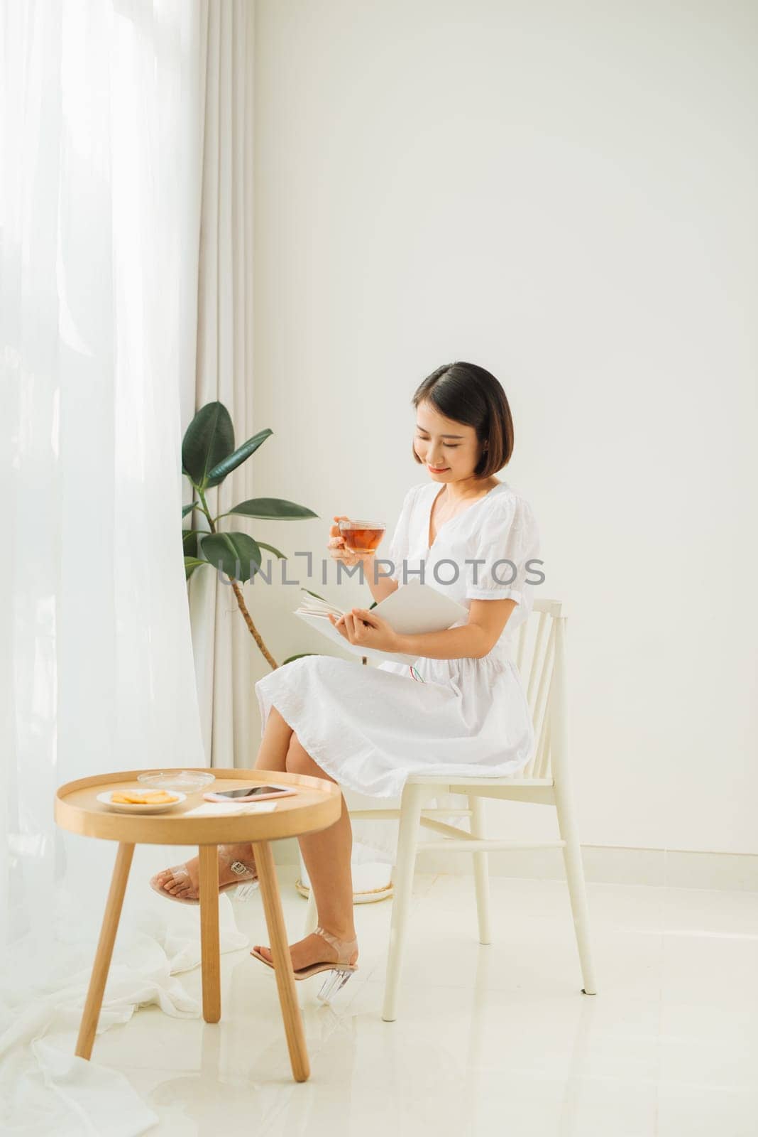 Young woman at home sitting near window relaxing in her living room reading book and drinking tea