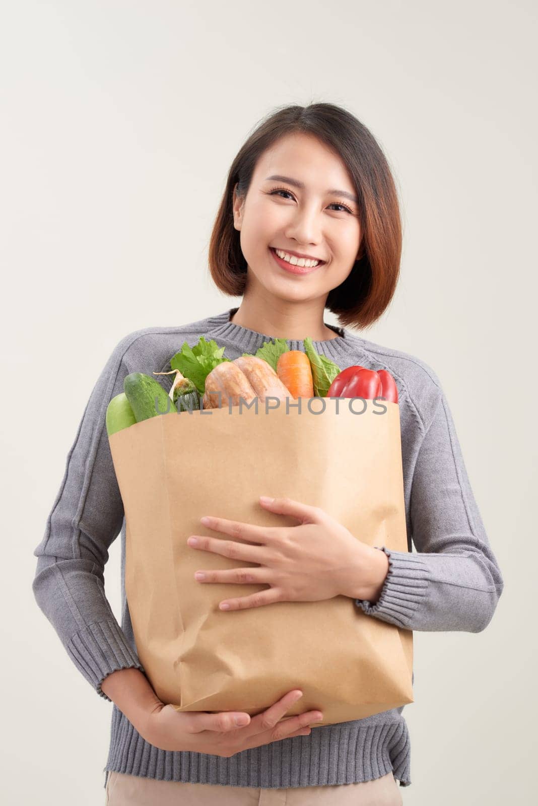 Beautiful smiling Asian woman holding paper shopping bag full of vegetables and groceries, studio shot isolated on white background by makidotvn