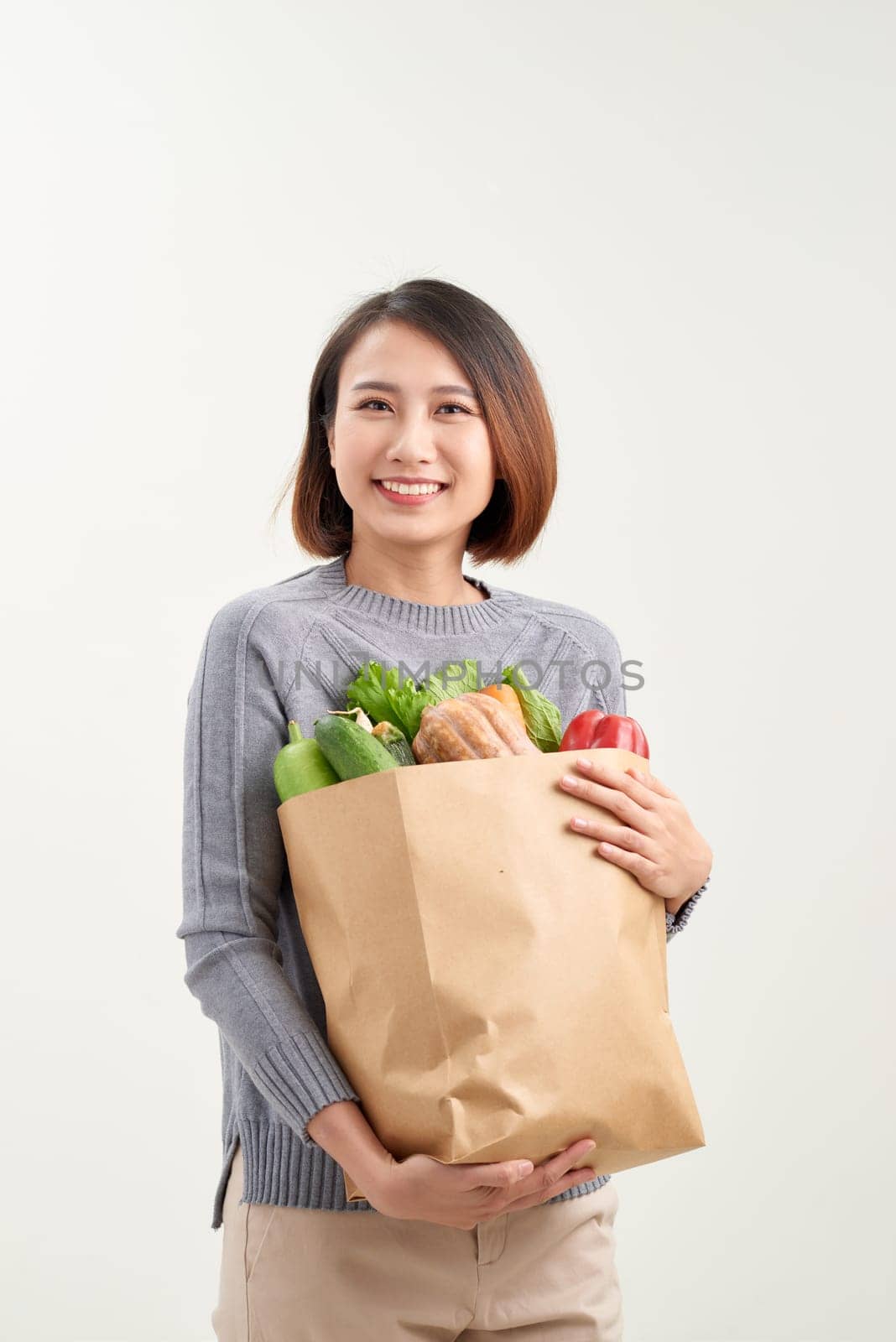 Beautiful young woman in apron holding paper shopping bag full of fresh vegetables and smiling