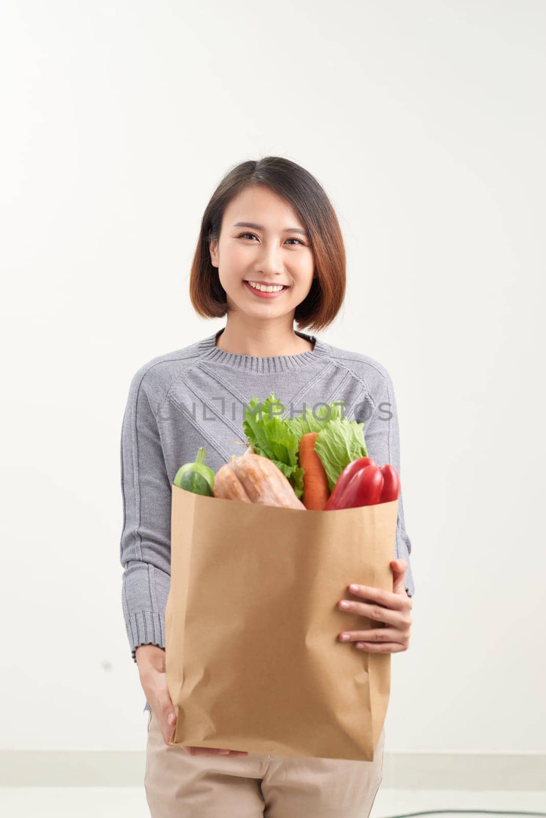 Beautiful smiling Asian woman holding paper shopping bag full of vegetables and groceries, studio shot isolated on white background