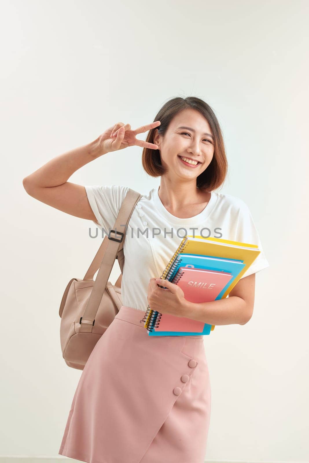 Cheerful pupil being in good mood after passing exam, shows with gesture everything is excellent, holds colourful folders  isolated over white background by makidotvn