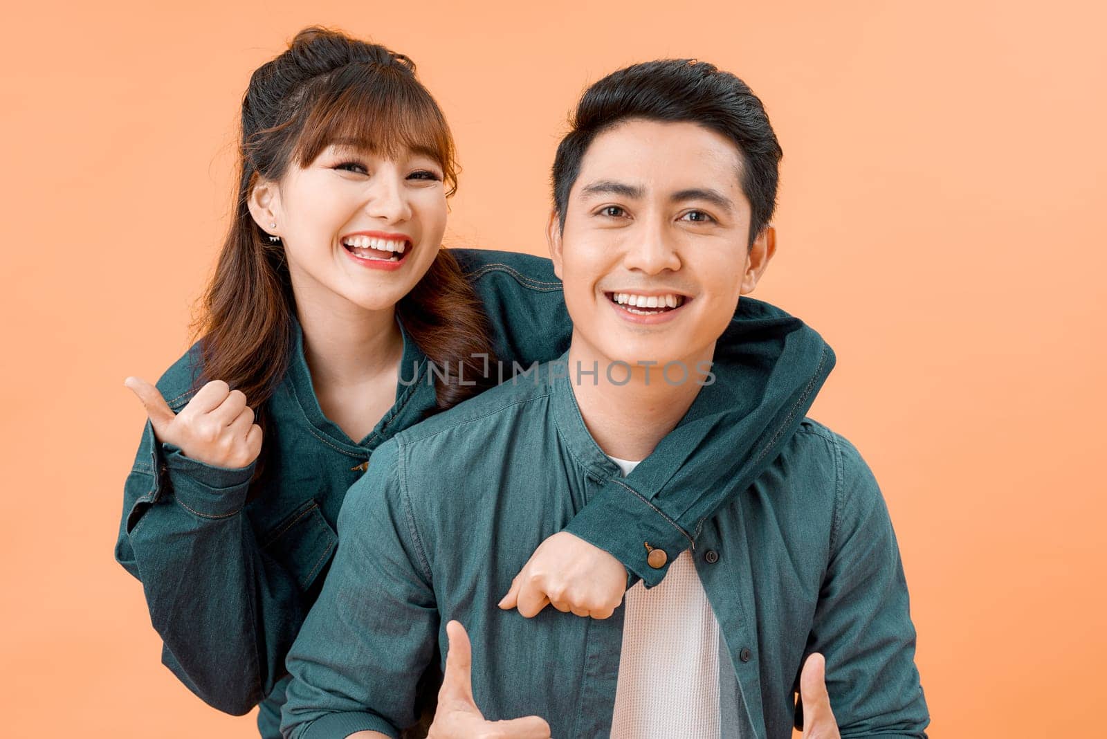 Portrait of a happy young couple hugging while standing and showing thumbs up gesture over yellow wall