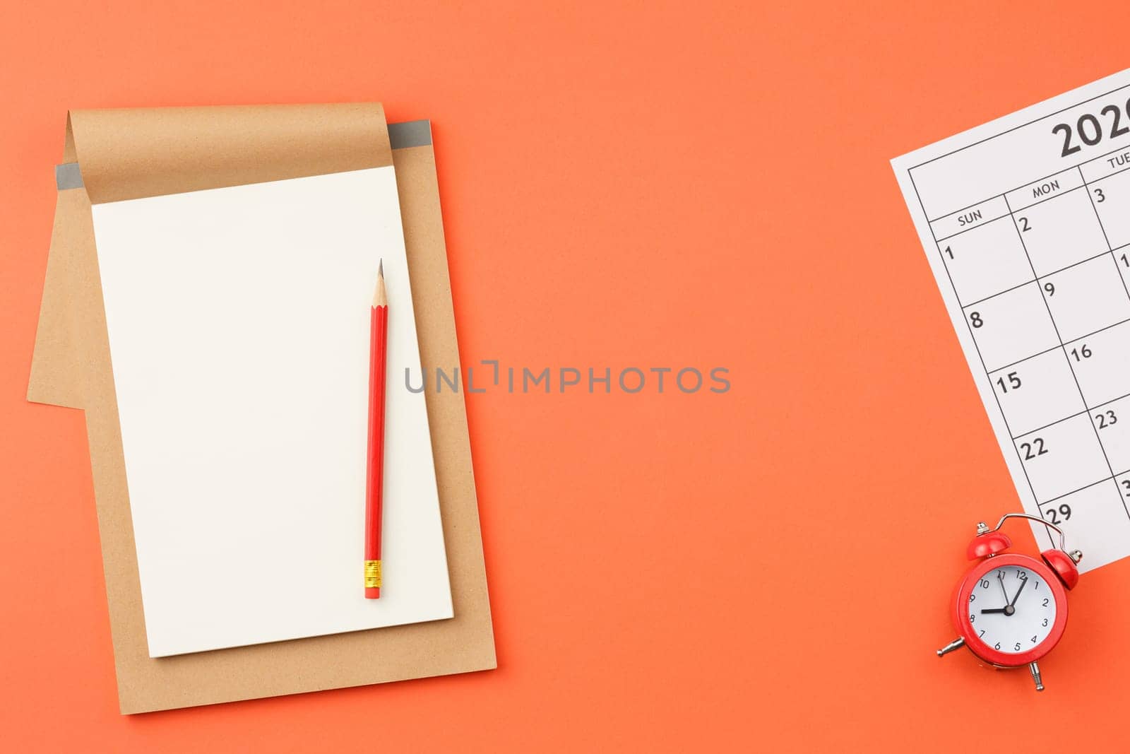 Craft notebook with white sheets, red alarm clock, pencil, and calendar sheet with dates on orange isolated background top view.