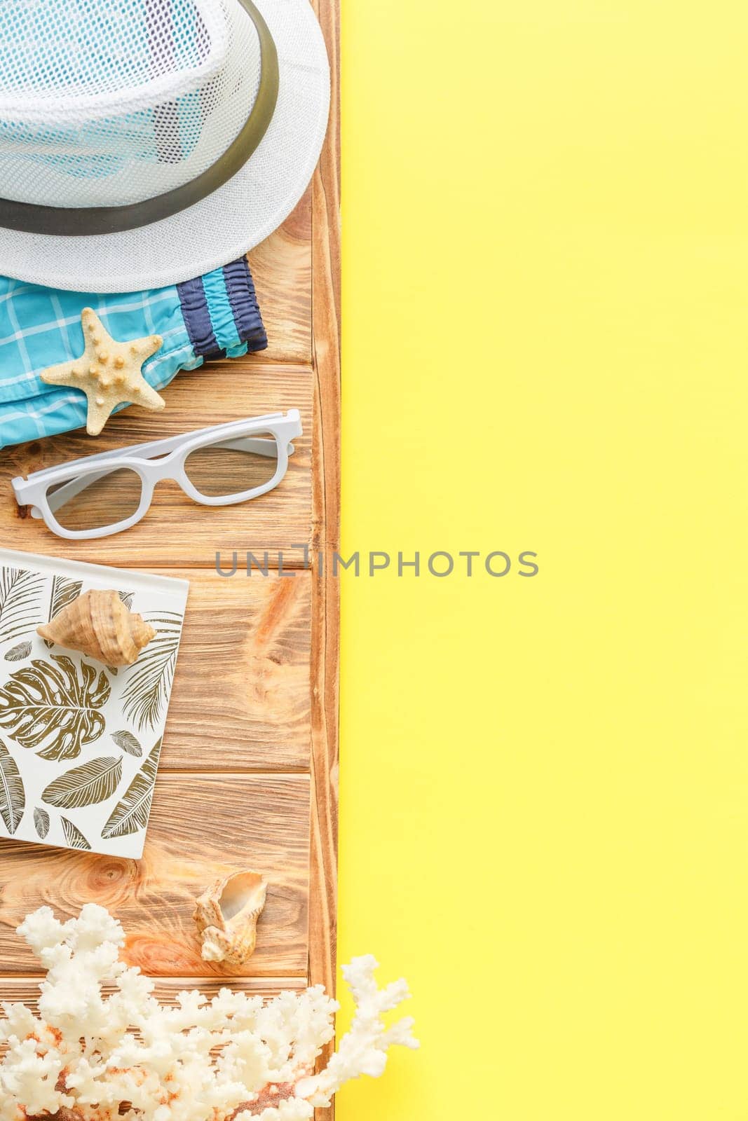 Beach hat with shorts and sunglasses on wooden background with yellow copy space. Flat lay. Summer vacation concept. Corals and seashell. Top view.