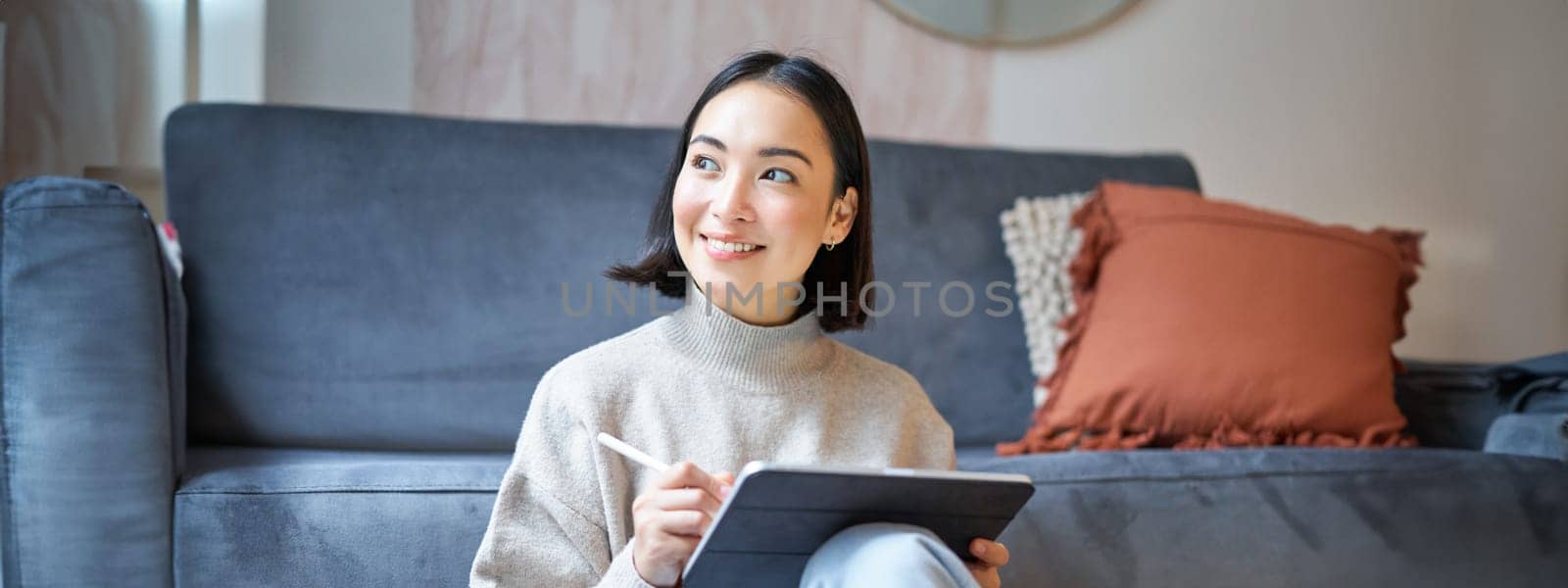 Portrait of asian woman with tablet, drawing, working on design project, holding pen, sitting in her living room, freelancing by Benzoix