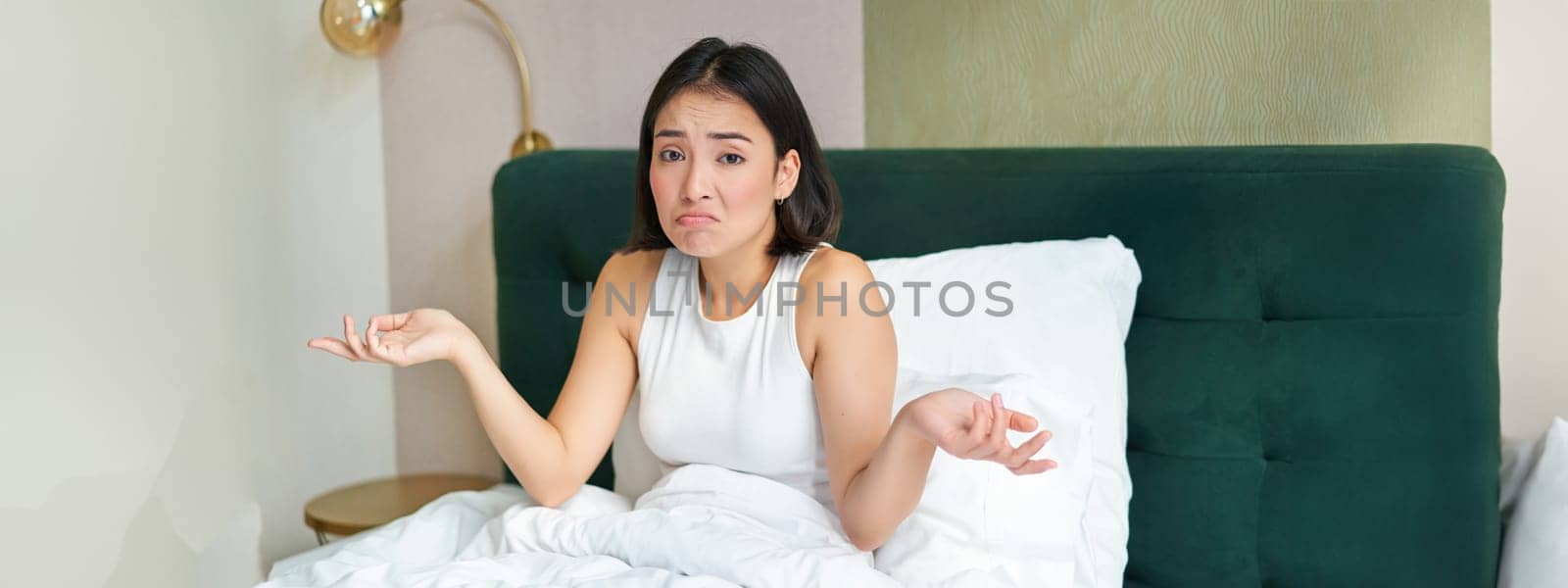 Confused asian girl lying in bed and shrugging shoulders, looking puzzled, dont know, spending time in her bedroom by Benzoix