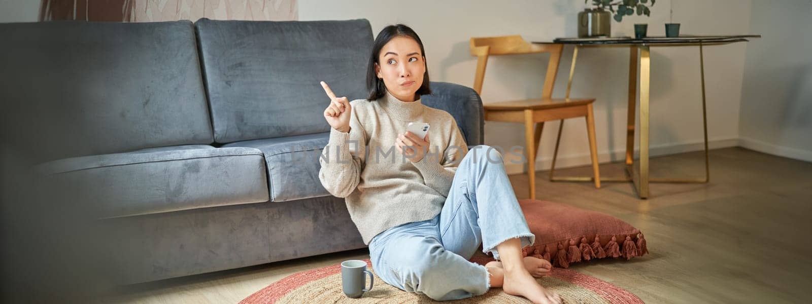 Portrait of woman sitting on floor with smartphone, looking thoughtful and pointing finger at banner, promo advertisement on top right corner.