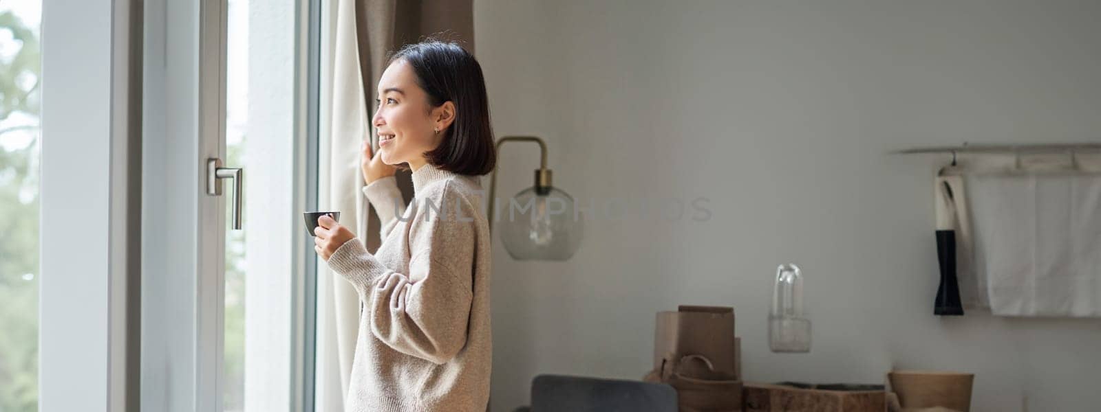 Beautiful korean woman staying at home, looking outside window, drinking coffee espresso and smiling, feeling comfort and warmth by Benzoix