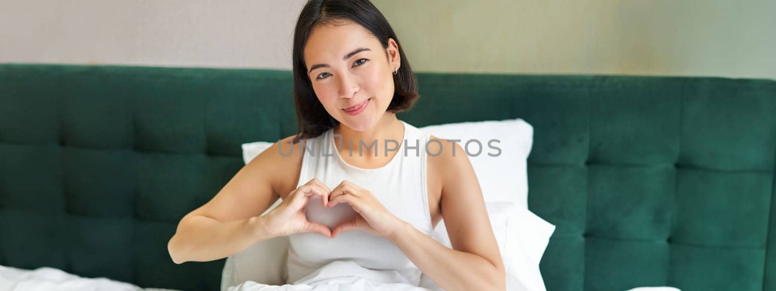 Tender asian girl shows heart sign, lying in bed, spending time in bedroom, expresses love and care.