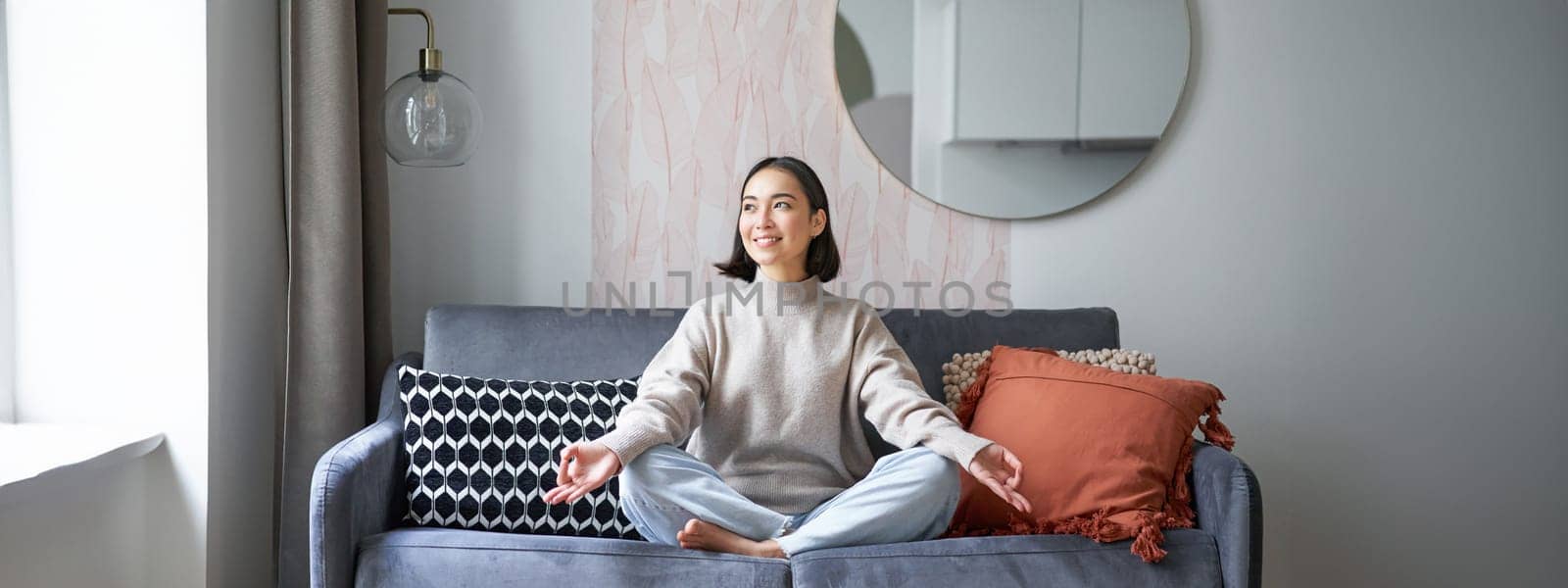 Relaxation and patience. Smiling young asian woman in cozy room, sitting on sofa and meditating, doing yoga mindfulness training.