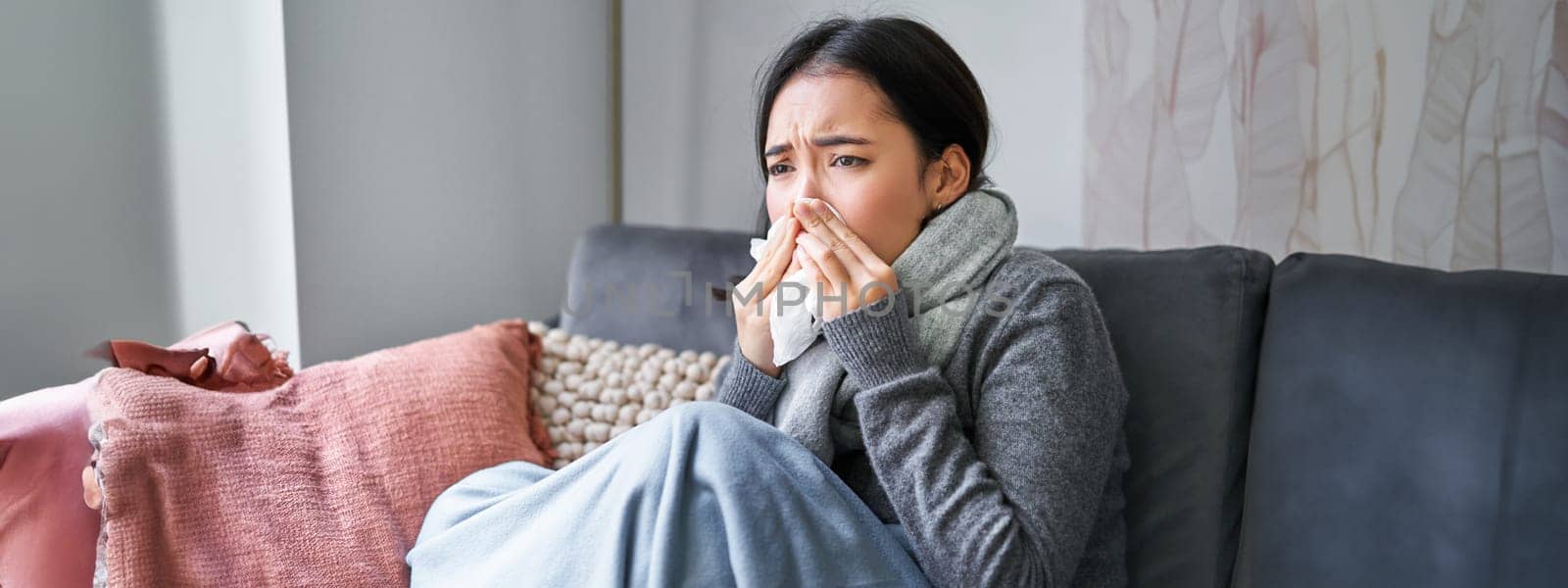 Image of sick korean woman at home, covered in warm clothes and scarf, feeling sick, catching a cold and sitting on sofa sneezing.