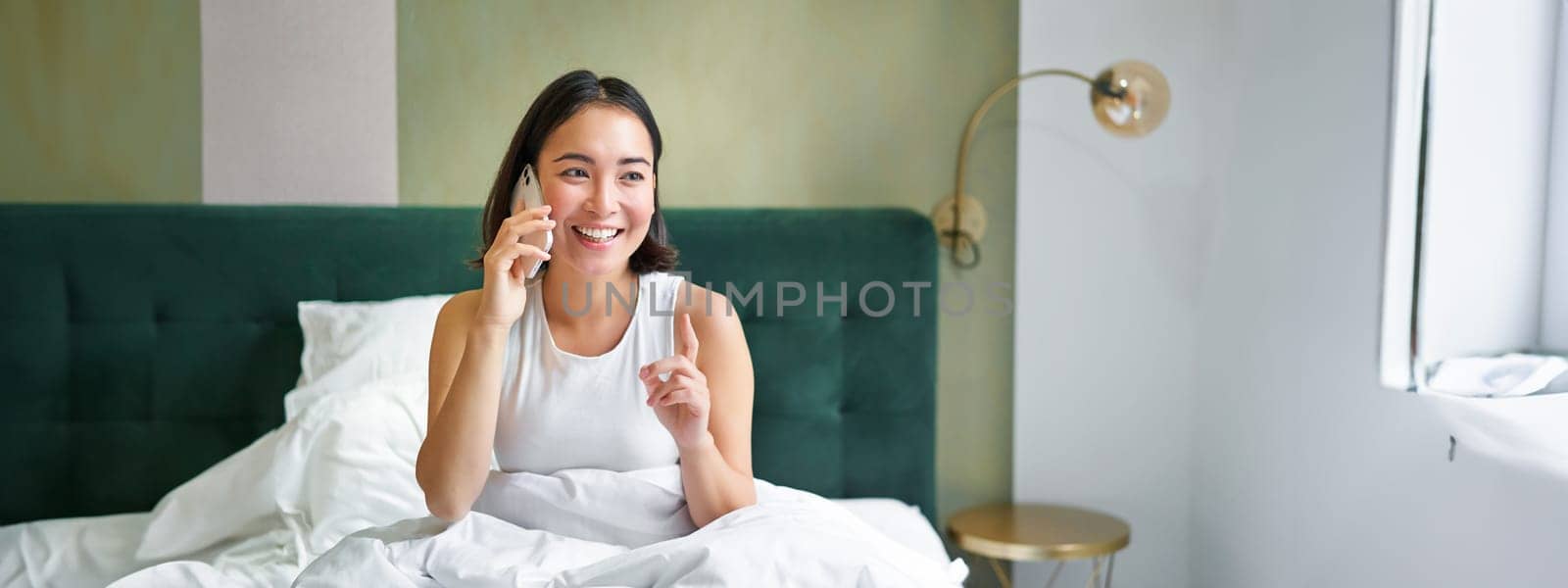 Beautiful smiling asian girl talking on mobile phone, lying in bed with pleased happy face, speaking to someone on telephone.