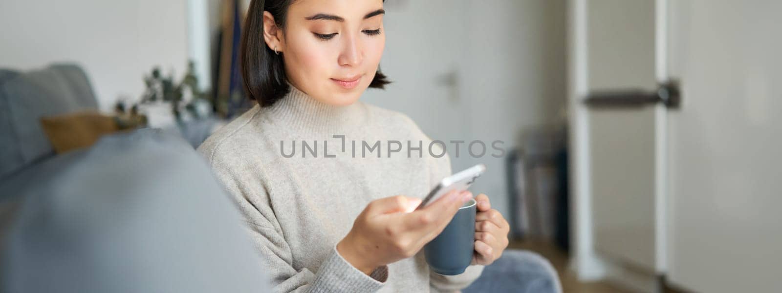 Portrait of smiling asian girl checking her news feed on smartphone and drinking coffee, sitting on sofa at home, browsing on mobile phone, reading.