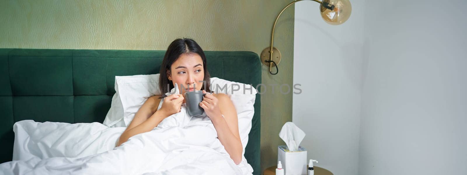 Health and people. Asian girl lying in bed, feeling sick, sneezing and drinking hot tea, catching cold, staying at home on leave.