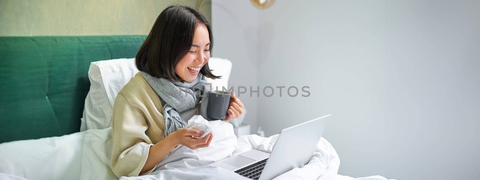 Portrait of asian sick girl, lying in bed with cold, having flu, talking to laptop, video chat, working from home on remote while on sick leave.