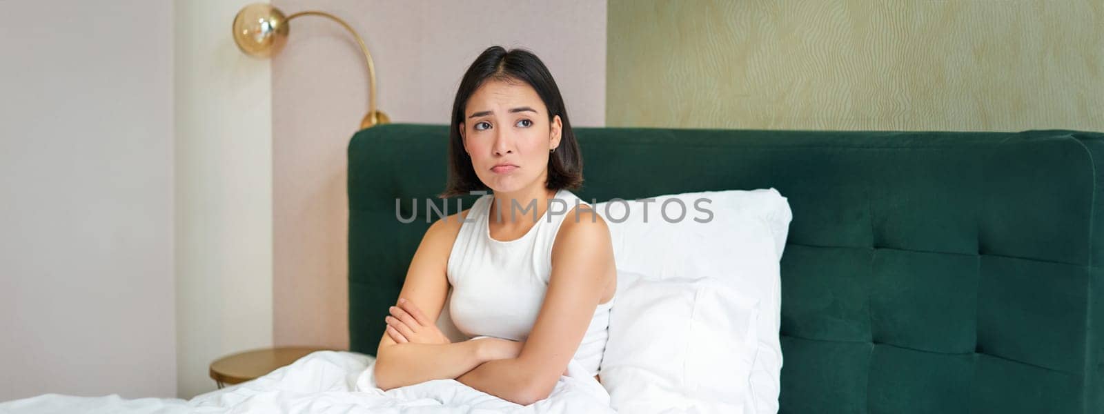 Portrait of asian girl in bed, looking complicated and worried, cross arms on chest, frowning with uneasy, thinking face expression.