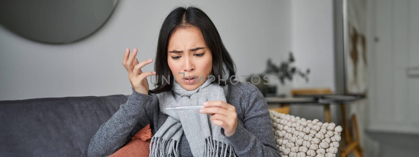 Shocked woman looks concerned at her thermometer, catching cold, has fever, sitting on sick leave at home by Benzoix