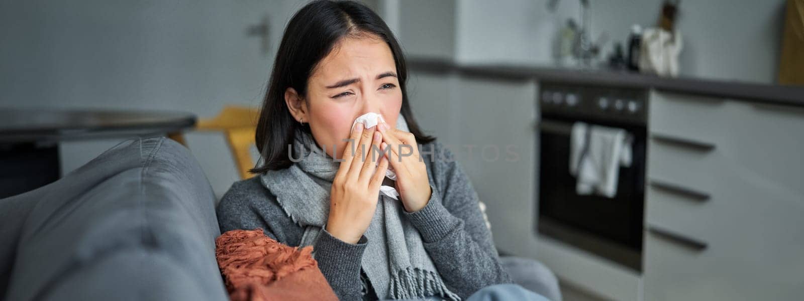 Portrait of ill young korean woman feeling sick, sneezing and holding napkin, staying at home ill, caught cold.