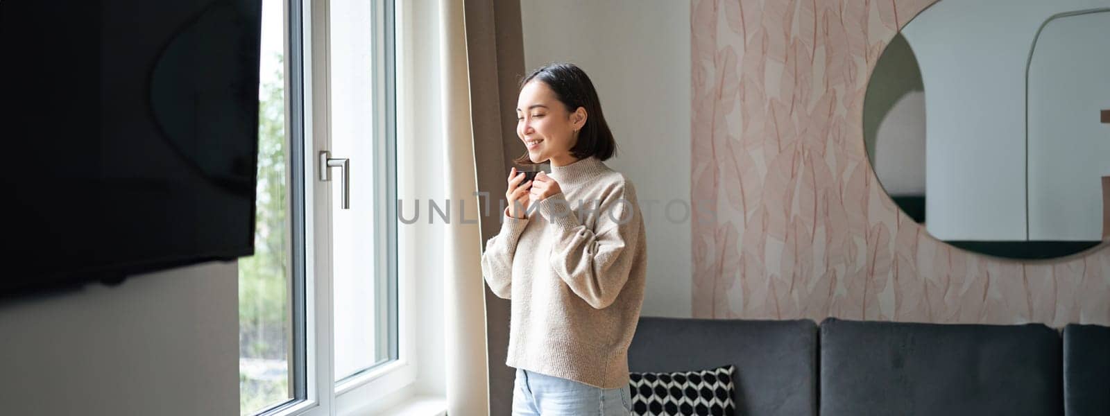 Morning. Happy asian woman drinking espresso at home, looking outside window and smiling, standing in cozy living room feeling warm.