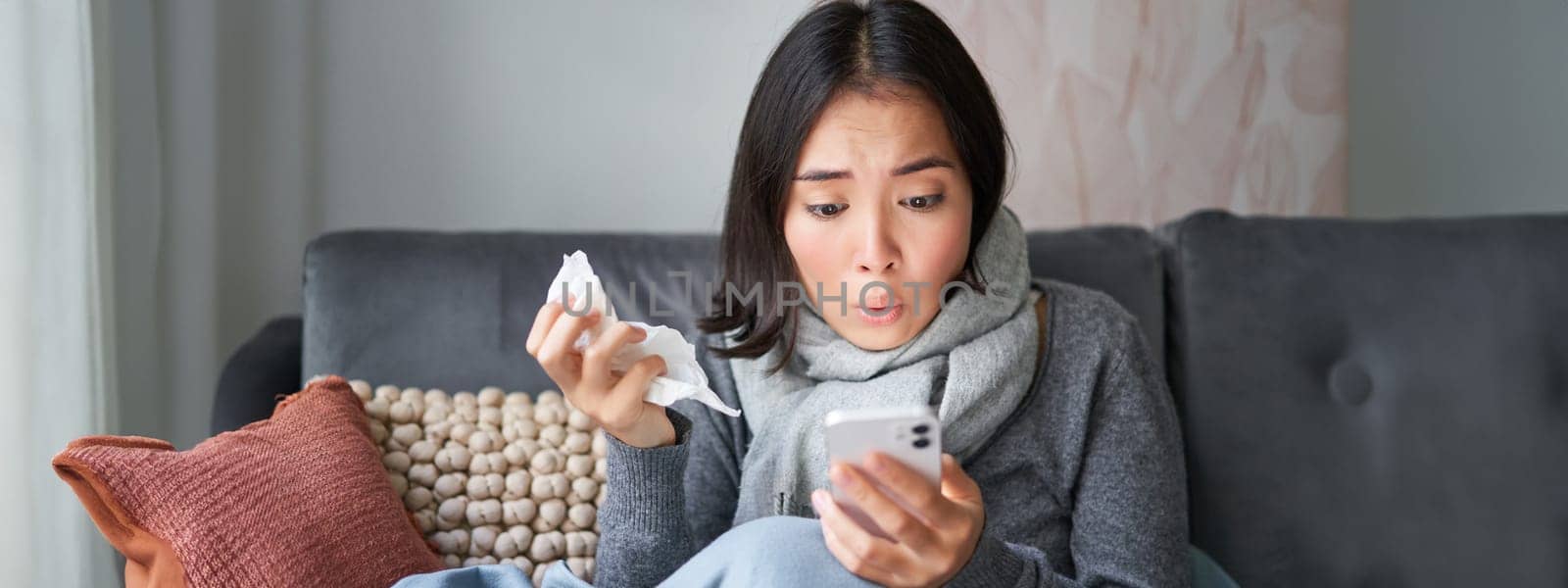 Portrait of ill woman looking at smartphone, looking concerned and shocked at mobile phone, reading bad news while sitting on sick leave at home.