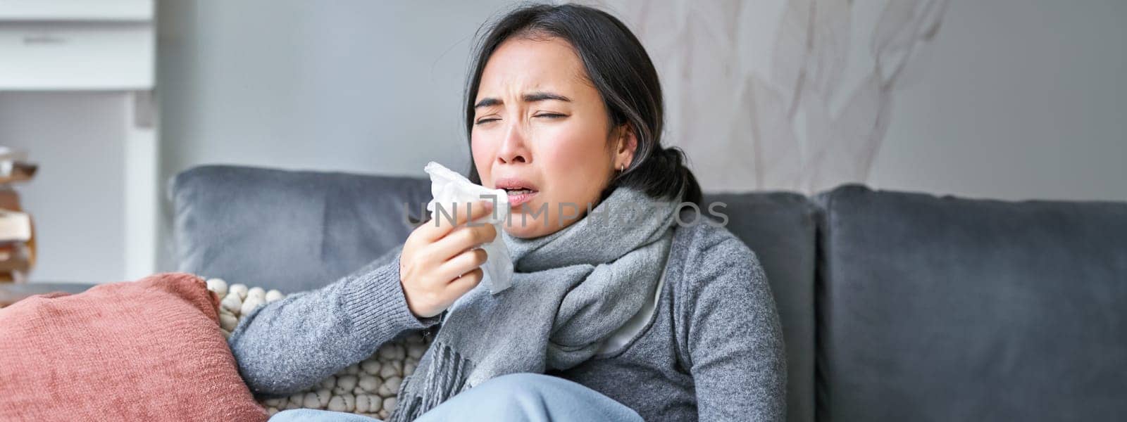 Close up of korean girl sneezing in napking, sitting at home in scarf, feeling unwell, concept of winter, heating cost and people.