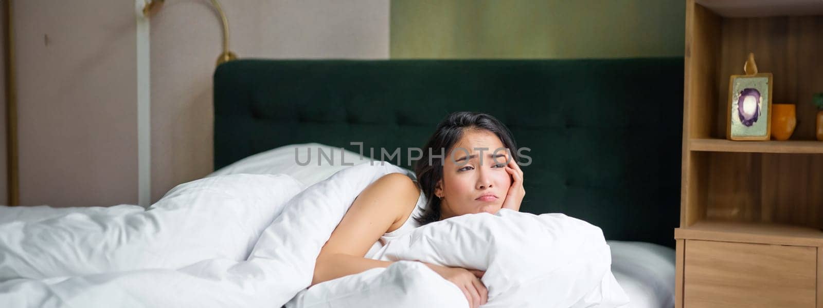 Portrait of upset korean woman, asian girl lying in bed feels sad, covers herself with duvet, looks outside window with disappointed face expression.