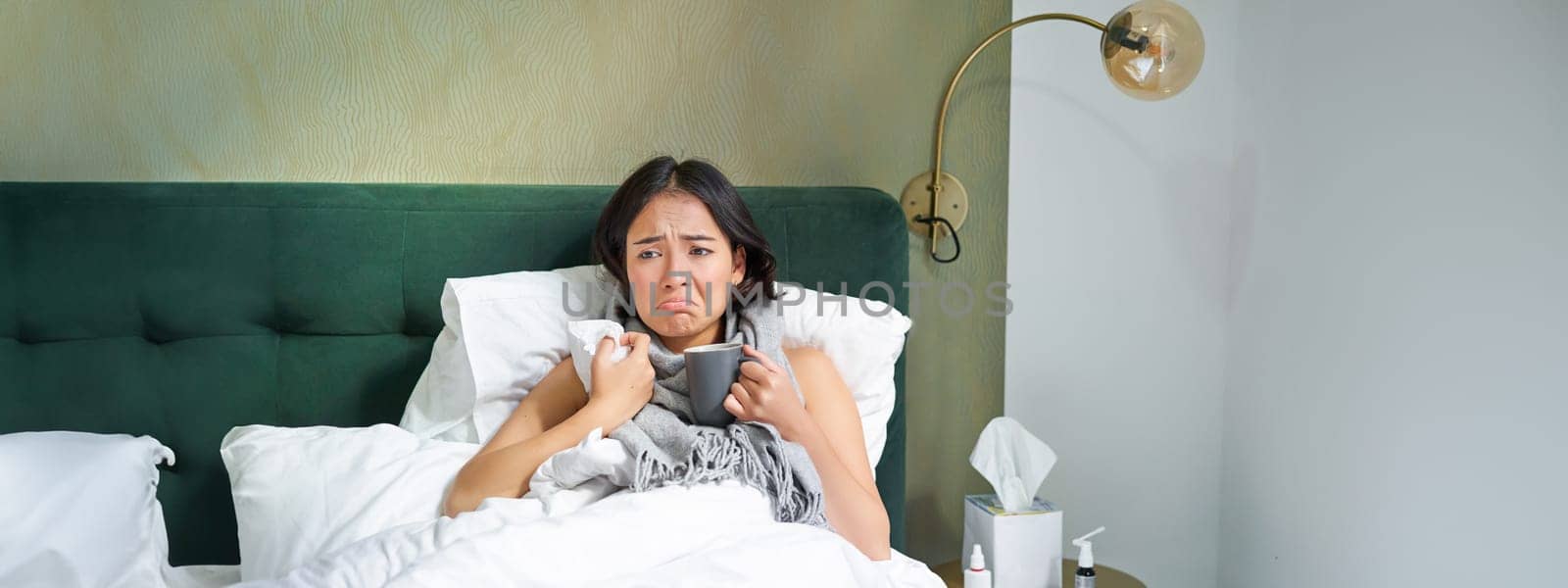 Sick sad asian girl lying in her bed with flu, catch a cold, taking pills and drinking hot tea, feeling upset.