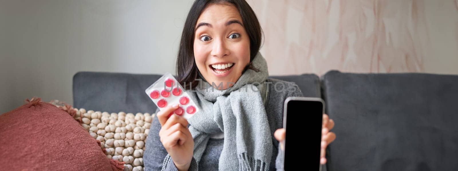 Smiling korean girl shows smartphone screen, medication in hands, feeling sick and staying at home, using online GP doctor app while on sick leave.