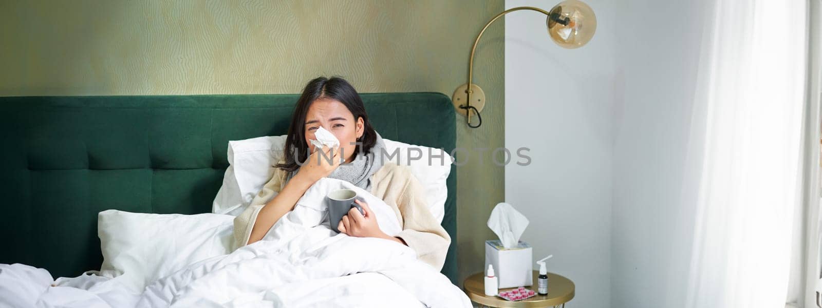 Covid-19, flu and vaccination concept. Korean girl lying in bed with cold, catching influenza, drinking hot tea and using prescribed medication by Benzoix