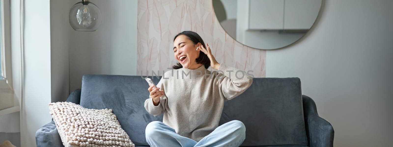 Portrait of carefree asian woman singing and listening music from smartphone app, using wireless headphones, smiling pleased, sitting on sofa at home.