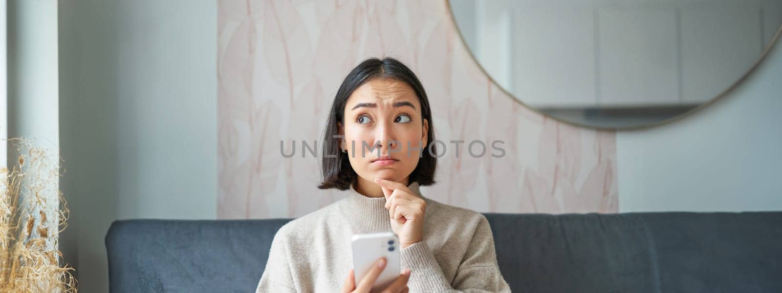 Portrait of girl sitting on sofa with smartphone, looking thoughtful and hesitant at mobile phone screen.