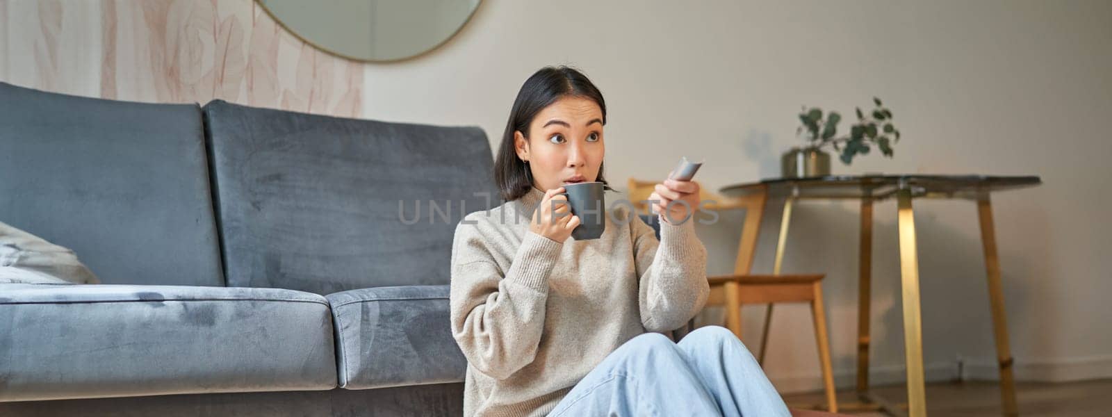 Portrait of young korean woman watching television, holding remote and looking amazed at tv screen, spending time at home.