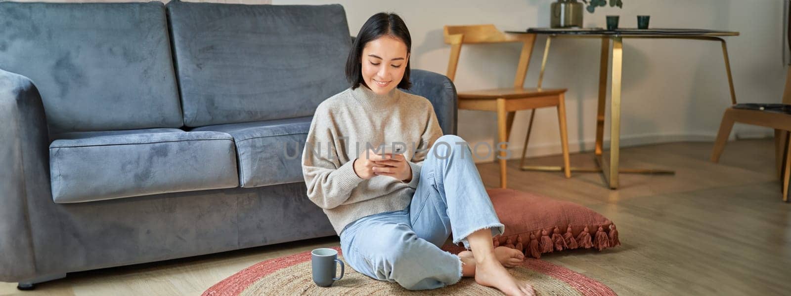 Image of stylish young woman in modern house, using mobile phone, sitting on floor and holding smartphone, drinking from cup.