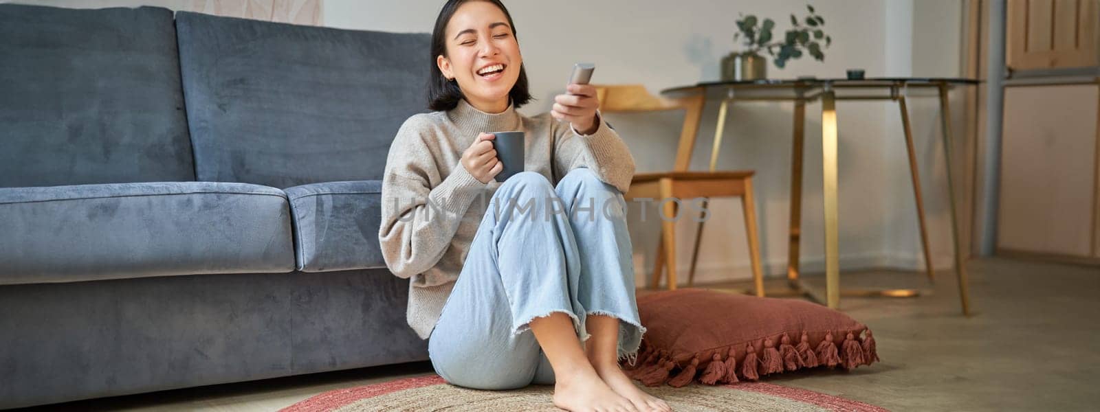 Portrait of young woman with remote, watching tv, switching chanels on television, sitting on floor near sofa and relaxing by Benzoix