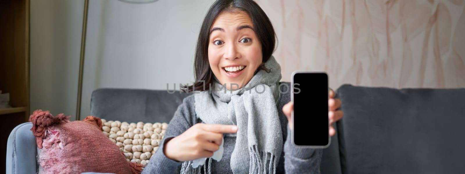 Excited young woman pointing finger at smartphone, showing online doctor, medical application or GP contact on mobile phone, staying at home sick, catching cold by Benzoix