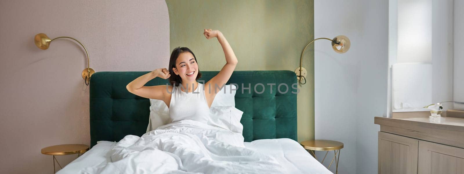 Beautiful asian woman lying in her bedroom on bed, stretching hands and looking outside, waking up from sleep.