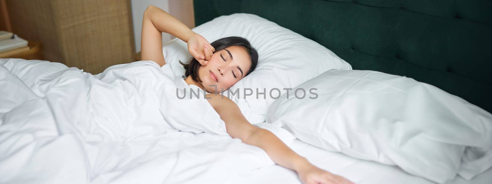 Romantic asian girl waking up in cozy bedroom, lying in bed with white sheets, stretching her hand towards empty pillow by Benzoix