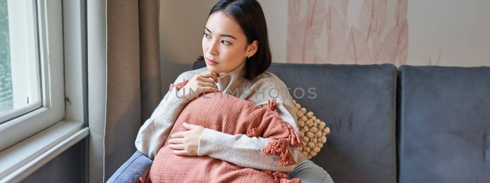 Portrait of asian woman feeling cozy in her home, hugging pillow on sofa and looking thoughtful, smiling.