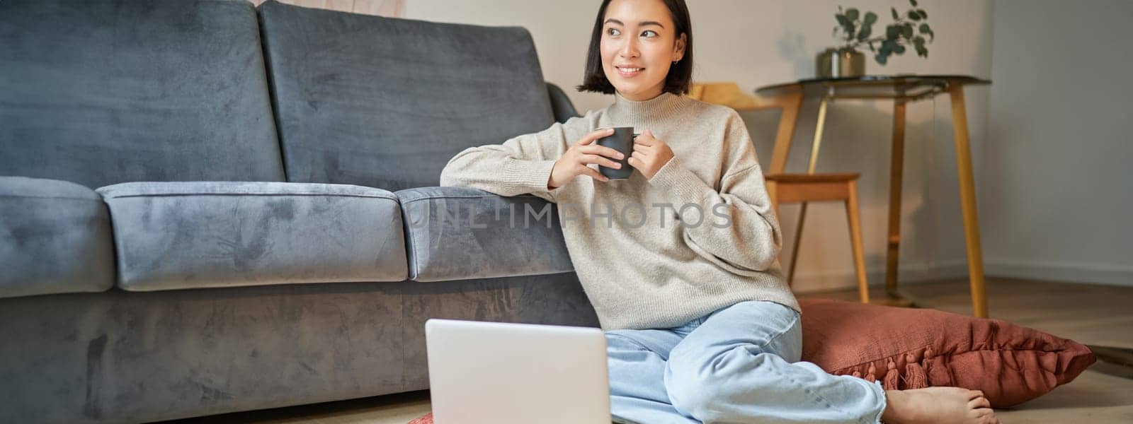 Image of smiling asian woman drinking hot tea, holding cup and sitting near laptop on floor, resting at home by Benzoix