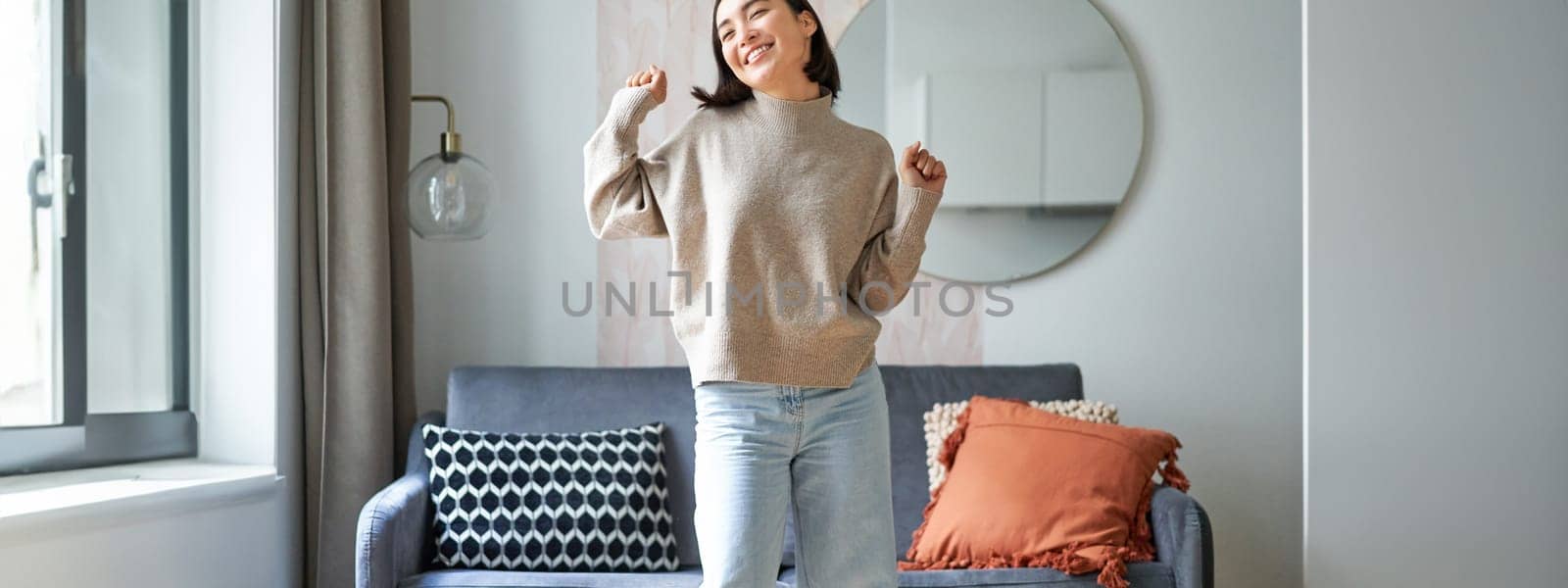 Enthusiastic asian woman dancing, feeling free and upbeat at her home, enjoying happy day, staying indoors, smiling and laughing.