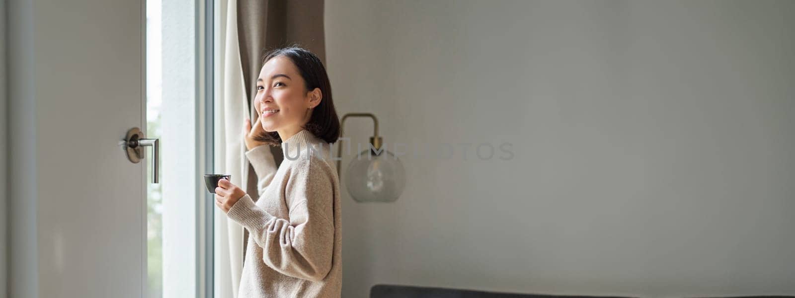 Beautiful korean woman staying at home, looking outside window, drinking coffee espresso and smiling, feeling comfort and warmth.