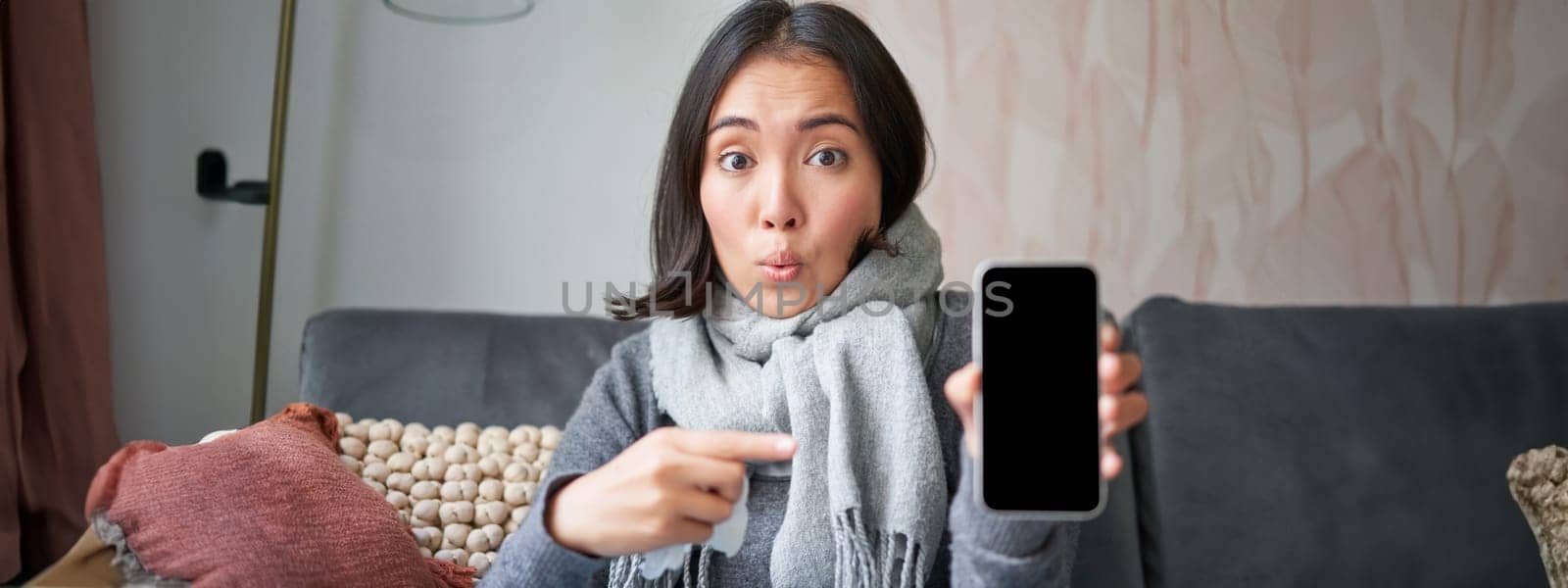 Excited young woman pointing finger at smartphone, showing online doctor, medical application or GP contact on mobile phone, staying at home sick, catching cold by Benzoix